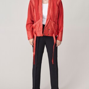 May include: A red leather jacket with a tie-front closure and long sleeves. The jacket has a relaxed fit and features a collar, pockets, and a belt.