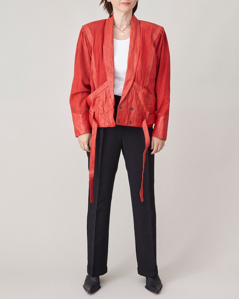 May include: A red leather jacket with a tie-front closure and long sleeves. The jacket has a relaxed fit and features a collar, pockets, and a belt.