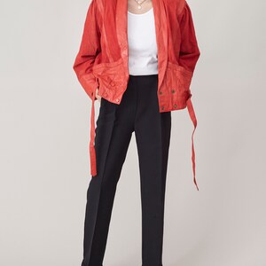 May include: A red leather jacket with a long, flowing tie at the waist. The jacket has a collar and a button closure. The model is wearing black pants and a white t-shirt.