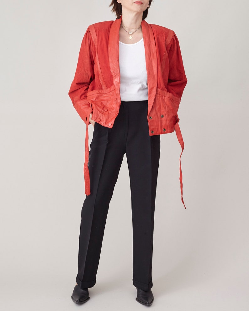 May include: A red leather jacket with a long, flowing tie at the waist. The jacket has a collar and a button closure. The model is wearing black pants and a white t-shirt.