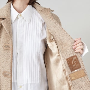 May include: A beige wool coat with a brown lining and a brown leather patch with the text "G.G.G. Garments" and "Sartorially Yours" on it.