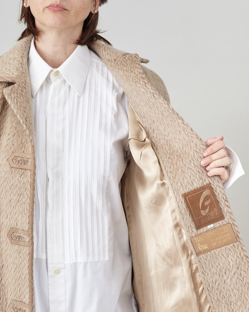 May include: A beige wool coat with a brown lining and a brown leather patch with the text "G.G.G. Garments" and "Sartorially Yours" on it.