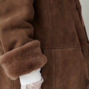 K&ouml;nnte beinhalten: Ein brauner Wildledermantel mit einem Pelzkragen und -&auml;rmelaufschl&auml;gen. Der Mantel hat einen Knopfverschluss und eine Tasche an der Seite.