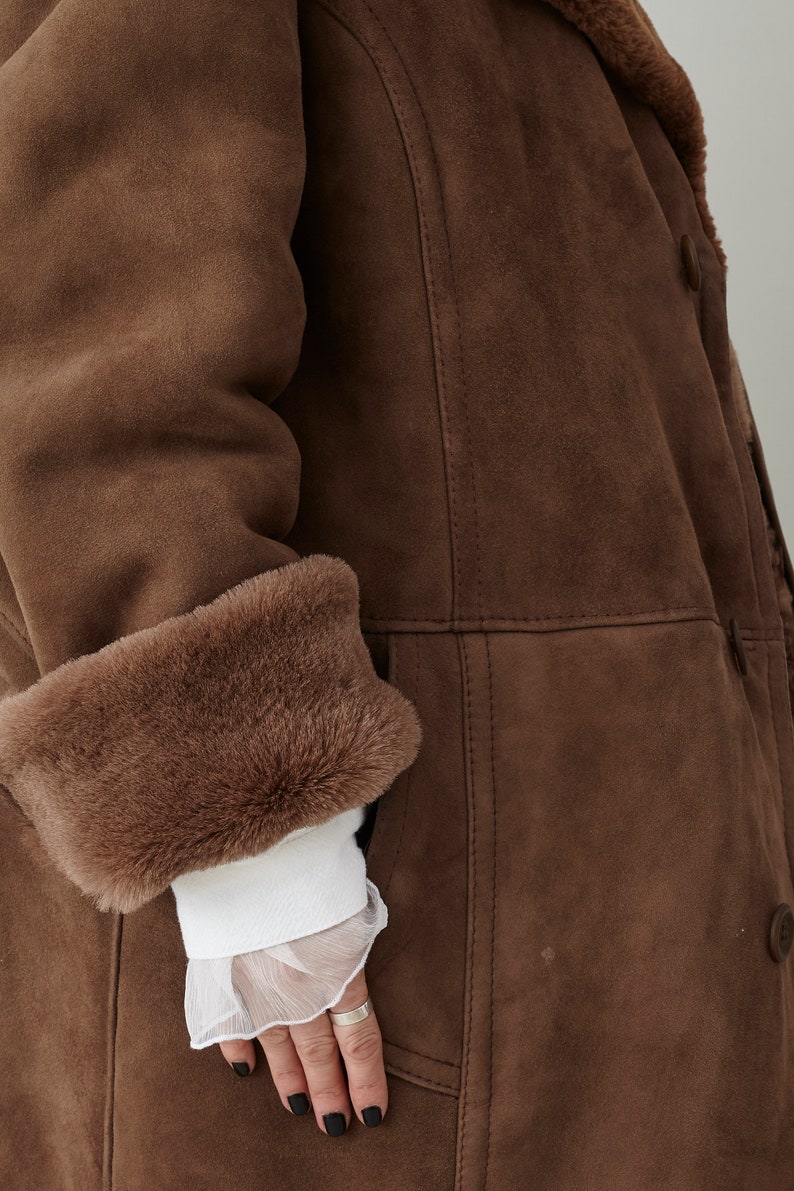 K&ouml;nnte beinhalten: Ein brauner Wildledermantel mit einem Pelzkragen und -&auml;rmelaufschl&auml;gen. Der Mantel hat einen Knopfverschluss und eine Tasche an der Seite.