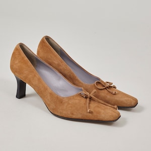 以下が含まれることがあります： スクエアトゥのブラウンのスエードのヒールシューズ。フロントにリボンがあしらわれています。