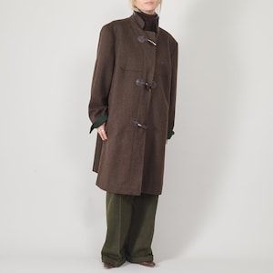 Peut inclure: Un manteau en laine marron avec des brandebourgs et un col montant. Le manteau a une forme évasée et une doublure verte aux poignets. Le modèle porte un pantalon vert et des chaussures marron. Vêtement d'extérieur classique.