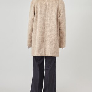 May include: A beige wool coat with a long, relaxed fit. The coat has a notched collar, a button closure, and long sleeves. The coat is shown from the back.