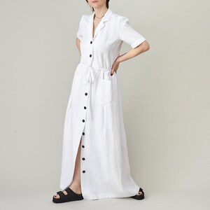 Puede incluir: Un vestido largo de lino blanco con botones en la parte delantera, mangas cortas y un lazo en la cintura.