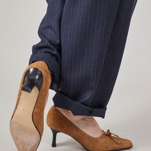 K&ouml;nnte beinhalten: Ein Paar braune Wildlederschuhe mit Absatz und einer Schleife vorne. Die Schuhe werden zu einer dunkelblauen Hose mit Nadelstreifen getragen.