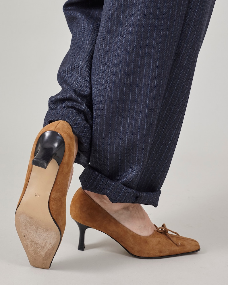 K&ouml;nnte beinhalten: Ein Paar braune Wildlederschuhe mit Absatz und einer Schleife vorne. Die Schuhe werden zu einer dunkelblauen Hose mit Nadelstreifen getragen.