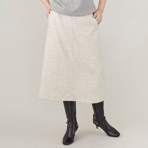 May include: A long, light gray skirt with a subtle herringbone pattern. The skirt is knee-length and has a straight silhouette. The model is wearing black knee-high boots.