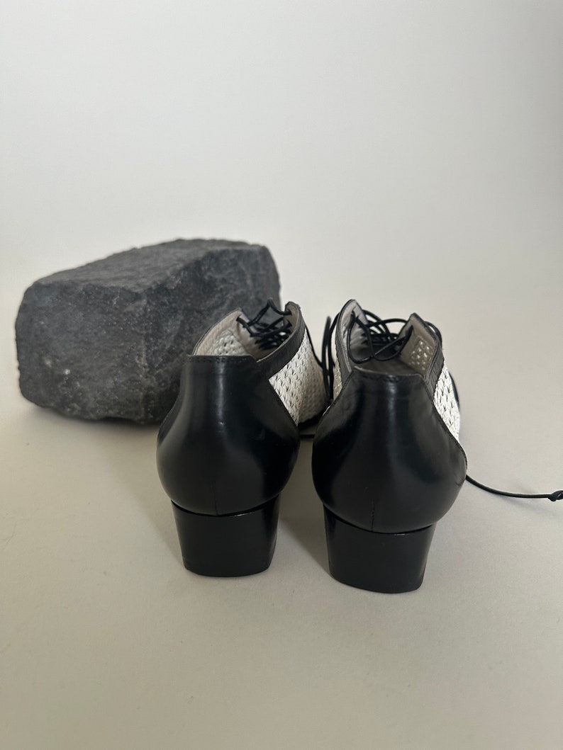 May include: A pair of black and white lace-up shoes with a low heel. The shoes have a woven pattern on the upper part and a solid black leather heel.