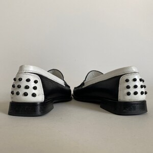 May include: Pair of black and white leather loafers. The shoes have a black body with a white heel and trim. The heel features a series of black studs. The shoes are displayed against a plain white background.