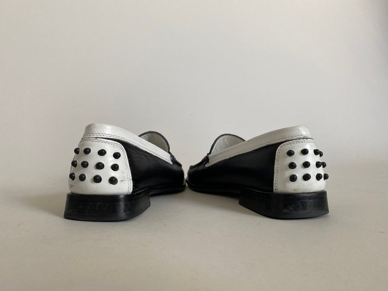 May include: Pair of black and white leather loafers. The shoes have a black body with a white heel and trim. The heel features a series of black studs. The shoes are displayed against a plain white background.