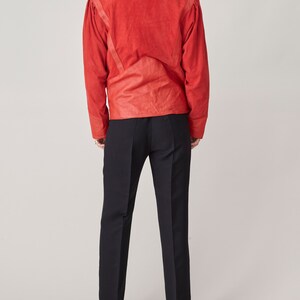 May include: A red suede jacket with a stand-up collar and long sleeves. The jacket has a fitted silhouette and features a unique design with seams and stitching details.