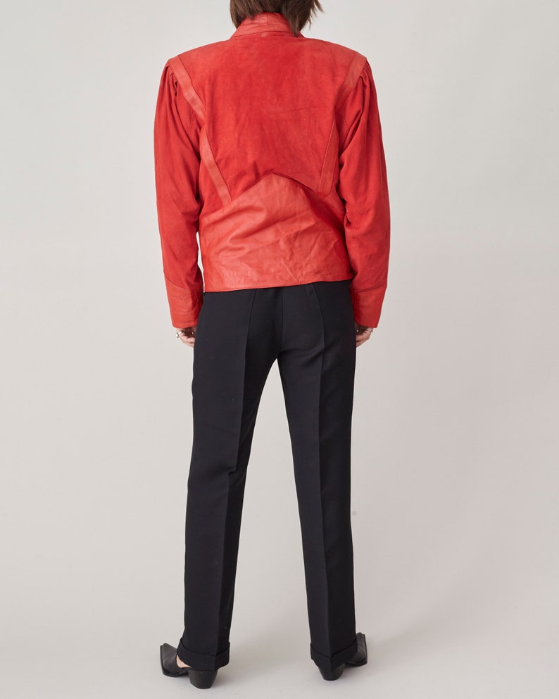 May include: A red suede jacket with a stand-up collar and long sleeves. The jacket has a fitted silhouette and features a unique design with seams and stitching details.