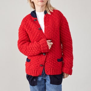 Könnte beinhalten: Ein leuchtend roter, grob gestrickter Cardigan mit marineblauen Verzierungen und Knöpfen. Der Cardigan hat eine strukturierte Optik und verfügt über einen runden Ausschnitt und zwei Vordertaschen. Die Ärmel sind umgeschlagen und das Kleidungsstück ist geknöpft.