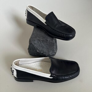 May include: Black leather loafers with white trim and accents. The shoes have a classic slip-on design with a stitched vamp and a rubber sole. The interior is white with a printed logo. The shoes are displayed on a dark gray rock.