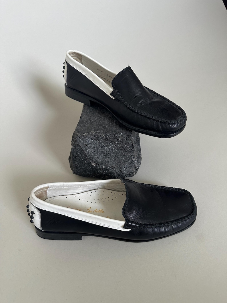 May include: Black leather loafers with white trim and accents. The shoes have a classic slip-on design with a stitched vamp and a rubber sole. The interior is white with a printed logo. The shoes are displayed on a dark gray rock.