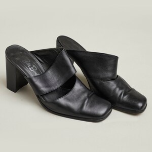 Puede incluir: Par de mules de tac&oacute;n de cuero negro con punta cuadrada y una sola tira en la parte superior. Los zapatos tienen un tac&oacute;n grueso y est&aacute;n etiquetados "Della Bedal" en la plantilla.