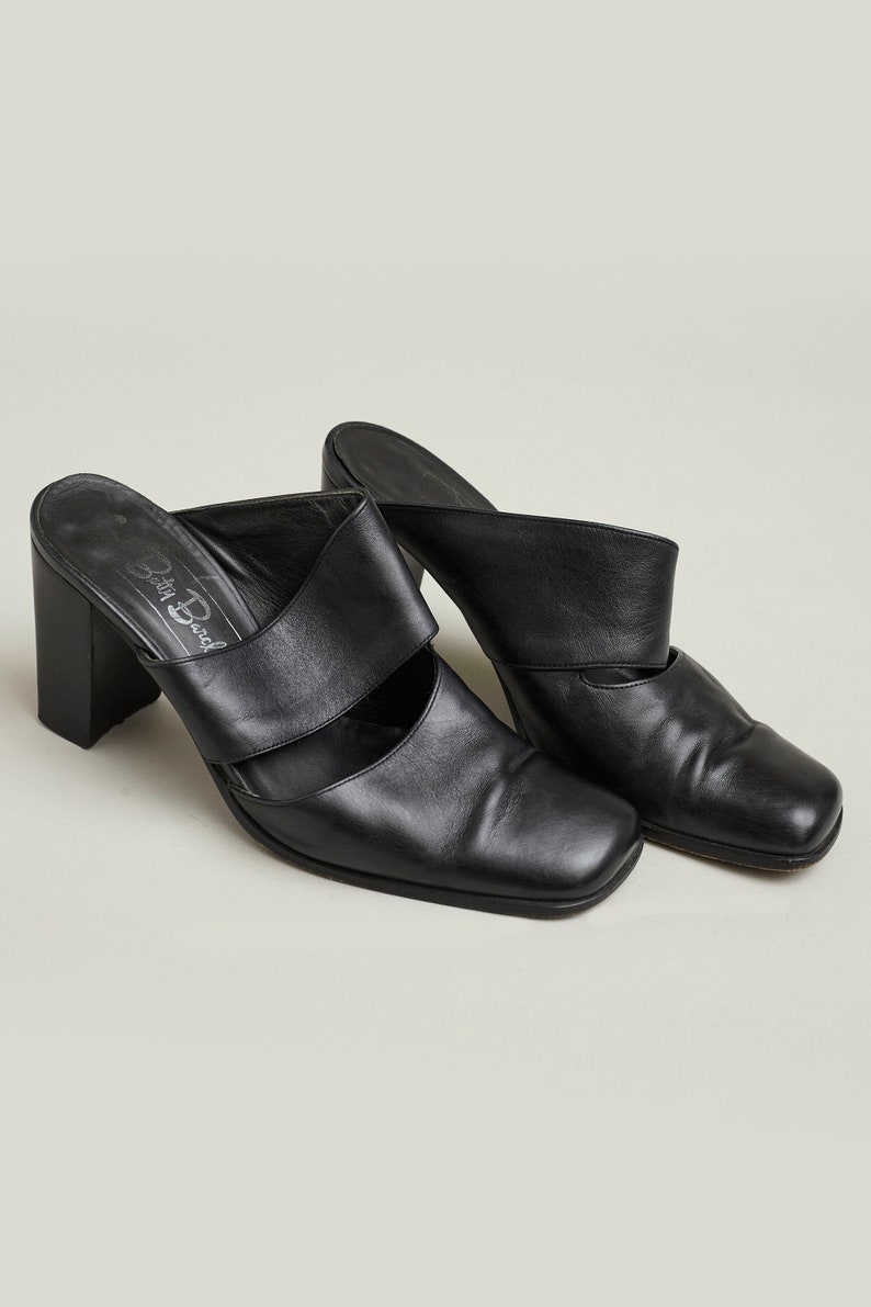 Puede incluir: Par de mules de tac&oacute;n de cuero negro con punta cuadrada y una sola tira en la parte superior. Los zapatos tienen un tac&oacute;n grueso y est&aacute;n etiquetados "Della Bedal" en la plantilla.