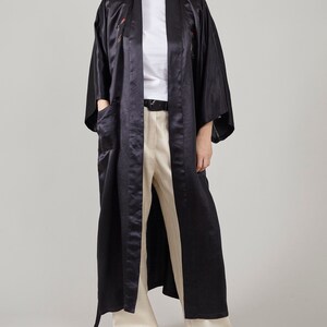 May include: A long black satin robe with a sash and embroidered details. The robe is worn over a white t-shirt and beige trousers.
