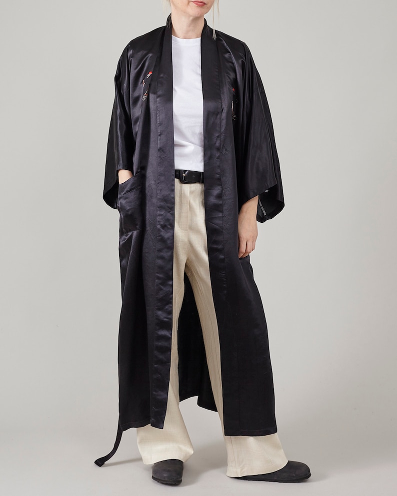 May include: A long black satin robe with a sash and embroidered details. The robe is worn over a white t-shirt and beige trousers.