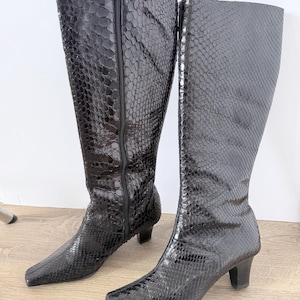 May include: A pair of knee-high boots in a glossy black finish with a textured snakeskin pattern. These boots feature a pointed toe, a modest block heel, and a full-length vertical zip closure along the inner side of the shaft for easy access.