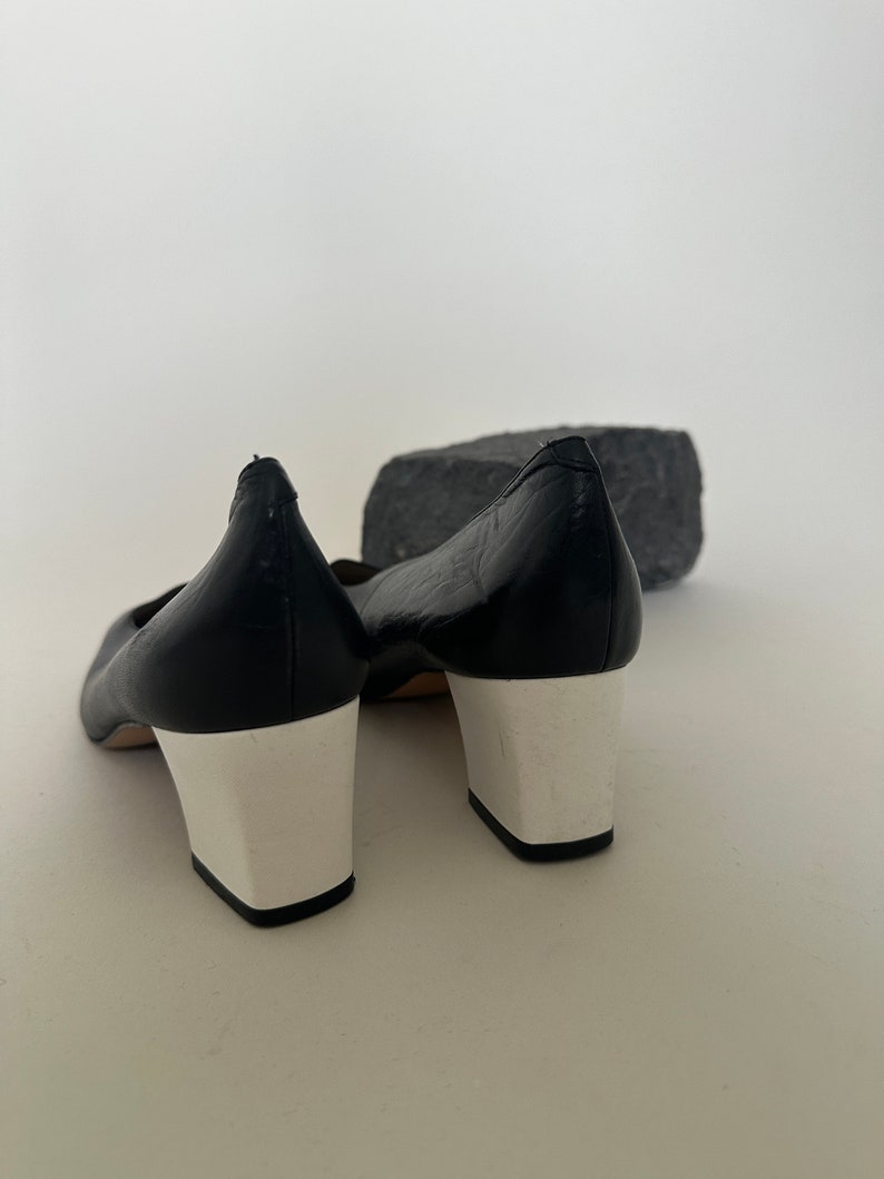 K&ouml;nnte beinhalten: Ein Paar schwarze Lederschuhe mit wei&szlig;en Abs&auml;tzen. Die Schuhe haben eine spitze Zehenpartie und einen niedrigen Absatz.
