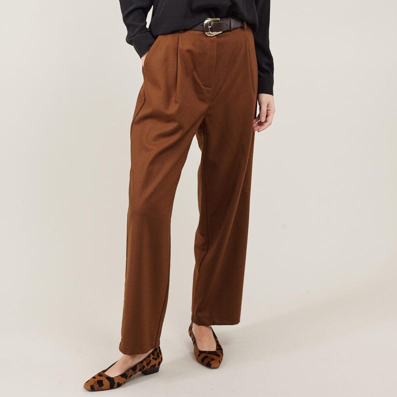 May include: Brown wide-leg pants with a black belt and a gold buckle. The pants have pleats at the front and are worn with brown and black leopard print flats.