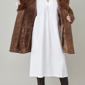 K&ouml;nnte beinhalten: Ein brauner Schaffellmantel mit einem Pelzkragen und einem wei&szlig;en Kleid darunter. Der Mantel ist offen und das Futter ist sichtbar. Die Person tr&auml;gt braune Stiefel.