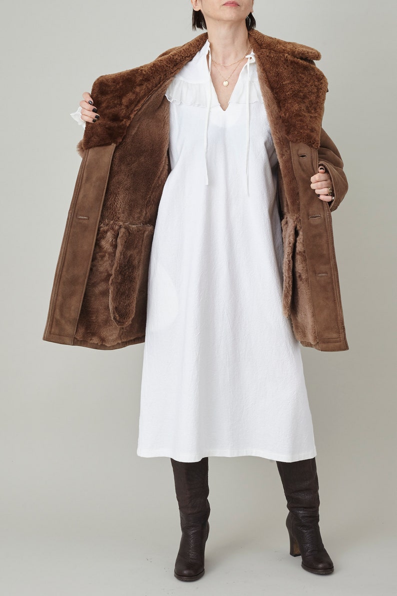 K&ouml;nnte beinhalten: Ein brauner Schaffellmantel mit einem Pelzkragen und einem wei&szlig;en Kleid darunter. Der Mantel ist offen und das Futter ist sichtbar. Die Person tr&auml;gt braune Stiefel.