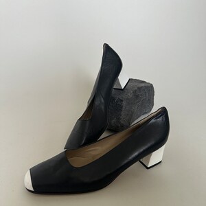 K&ouml;nnte beinhalten: Ein Paar schwarze Lederschuhe mit Absatz und wei&szlig;er Verzierung an Zehen und Absatz. Die Schuhe stehen auf einer wei&szlig;en Oberfl&auml;che.