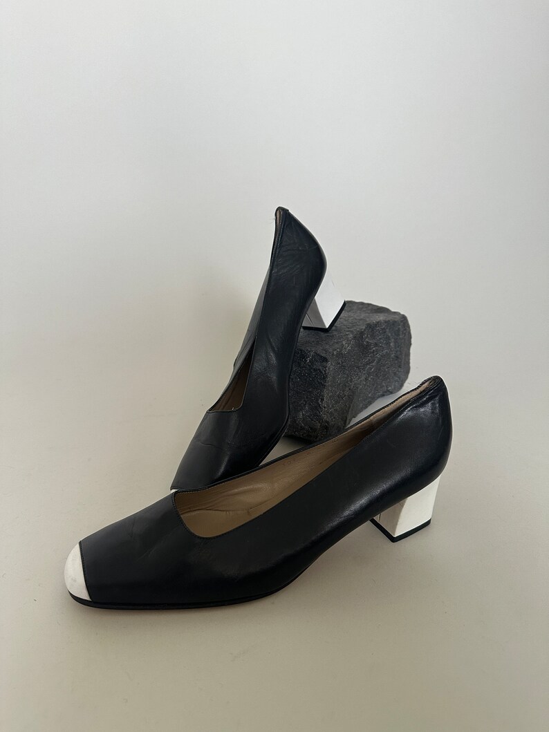 K&ouml;nnte beinhalten: Ein Paar schwarze Lederschuhe mit Absatz und wei&szlig;er Verzierung an Zehen und Absatz. Die Schuhe stehen auf einer wei&szlig;en Oberfl&auml;che.