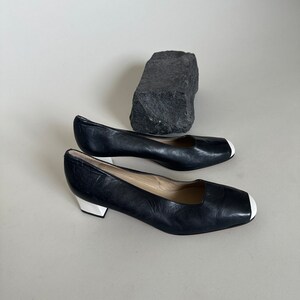 K&ouml;nnte beinhalten: Ein Paar schwarze Lederschuhe mit Absatz und wei&szlig;em Besatz an der Spitze. Die Schuhe befinden sich auf einem wei&szlig;en Hintergrund.