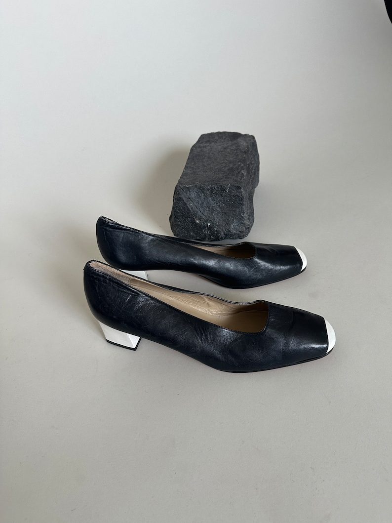K&ouml;nnte beinhalten: Ein Paar schwarze Lederschuhe mit Absatz und wei&szlig;em Besatz an der Spitze. Die Schuhe befinden sich auf einem wei&szlig;en Hintergrund.