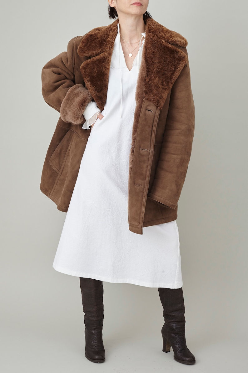 K&ouml;nnte beinhalten: Ein brauner Schaffellmantel mit einem Pelzkragen und einem wei&szlig;en Kleid darunter. Der Mantel wird von einer Person mit braunen Haaren und braunen Stiefeln getragen.