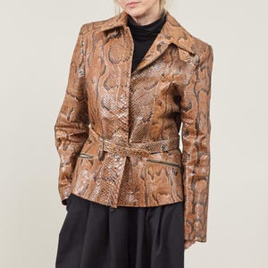 Pode incluir: Uma jaqueta de couro de cobra marrom com cintura cintada e fecho de zíper. A jaqueta tem gola e mangas compridas.