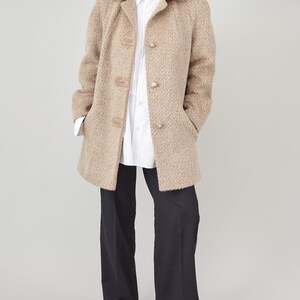 May include: A beige wool coat with a herringbone pattern and leather button accents. The coat has a collar and long sleeves. The model is wearing a white button-down shirt and black trousers.