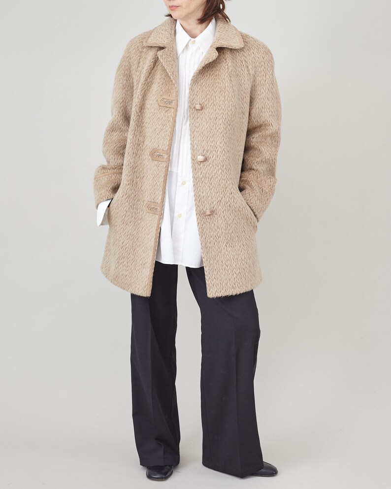May include: A beige wool coat with a herringbone pattern and leather button accents. The coat has a collar and long sleeves. The model is wearing a white button-down shirt and black trousers.