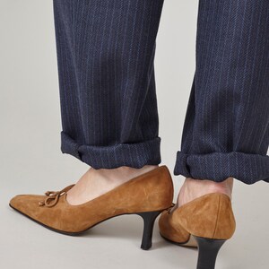 K&ouml;nnte beinhalten: Ein Paar braune Wildlederschuhe mit Absatz, spitzer Zehenpartie und Schleife vorne. Die Schuhe werden zu einer dunkelblauen Hose mit Nadelstreifen getragen.