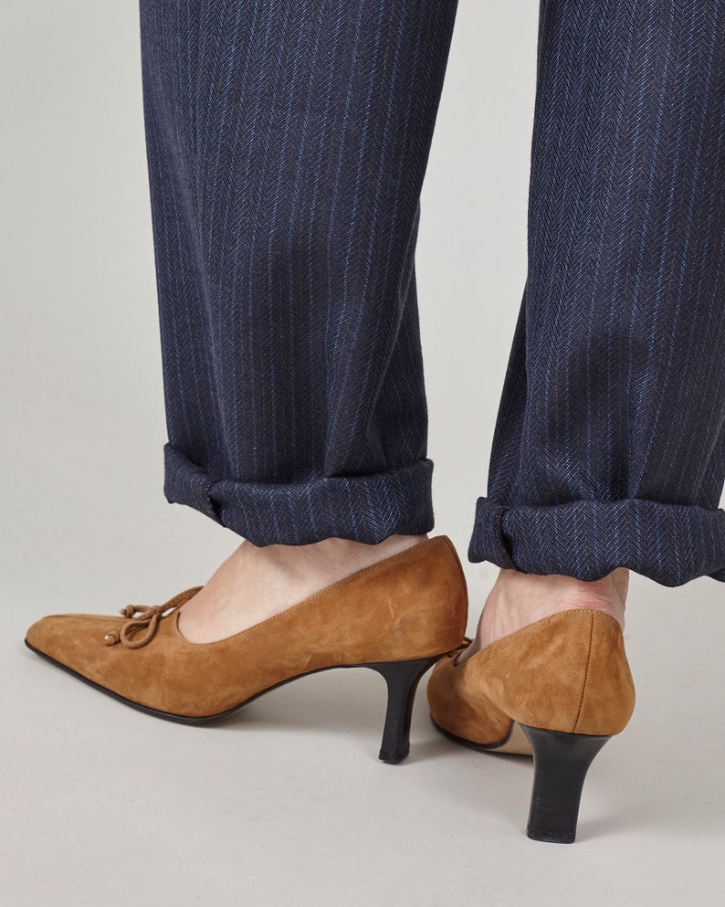 K&ouml;nnte beinhalten: Ein Paar braune Wildlederschuhe mit Absatz, spitzer Zehenpartie und Schleife vorne. Die Schuhe werden zu einer dunkelblauen Hose mit Nadelstreifen getragen.