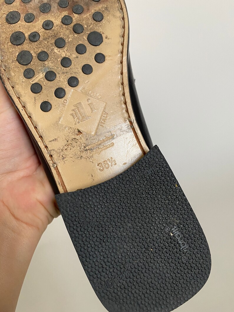May include: Close-up of the sole of a black leather shoe. The sole is worn, with a rubber heel and a textured rubber sole. The shoe size is 36 1/2. The sole has the words "Vero Cuoio Italy" and the brand "Vibram".