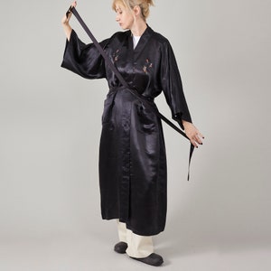 May include: A black satin robe with a long, flowing silhouette and a sash tie. The robe features embroidered birds and is worn by a person with blonde hair.