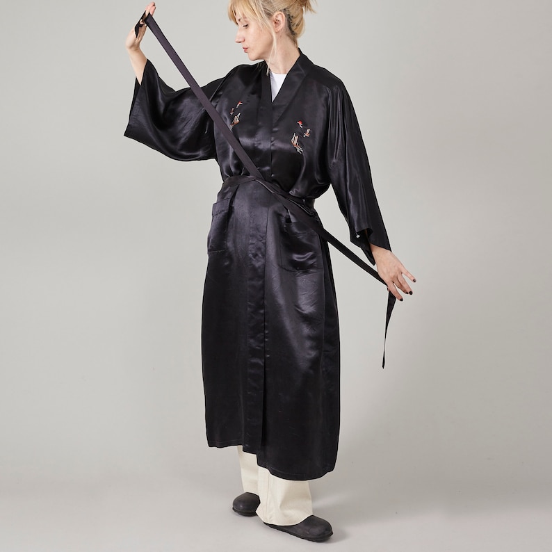 May include: A black satin robe with a long, flowing silhouette and a sash tie. The robe features embroidered birds and is worn by a person with blonde hair.