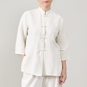 May include: A white linen shirt with a mandarin collar and five decorative buttons. The shirt has a relaxed fit and three-quarter sleeves.