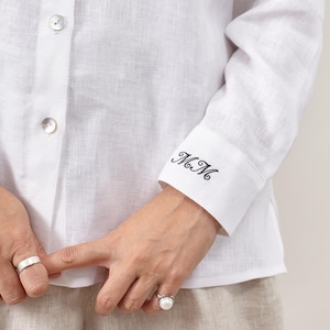 Puede incluir: Camisa de lino blanca con mangas largas y abotonadura. El puño de la manga está bordado con las iniciales "M.M" en escritura negra. La camisa se combina con pantalones de lino beige. Se ven anillos de plata.