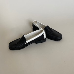 May include: A pair of black leather loafers with white trim. The shoes have a classic design with a rounded toe and a low heel. The interior of the shoe is white. The shoes are displayed on a neutral background.