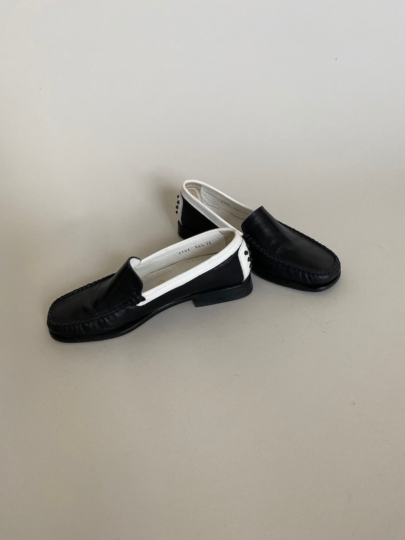 May include: A pair of black leather loafers with white trim. The shoes have a classic design with a rounded toe and a low heel. The interior of the shoe is white. The shoes are displayed on a neutral background.