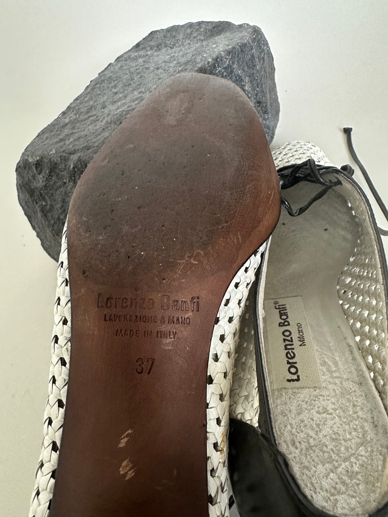 May include: White leather woven sandals with black leather trim and a brown leather sole. The sandals have a label that reads "Lorenzo Banfi" and "Lavorazione a Mano Made in Italy". The size is 37.